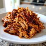 A close-up of tender, shredded crock pot pulled pork coated in rich barbecue sauce, served on a white plate.