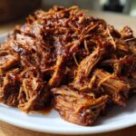 Close-up of tender, shredded crock pot pulled pork coated in rich barbecue sauce, served on a white plate.