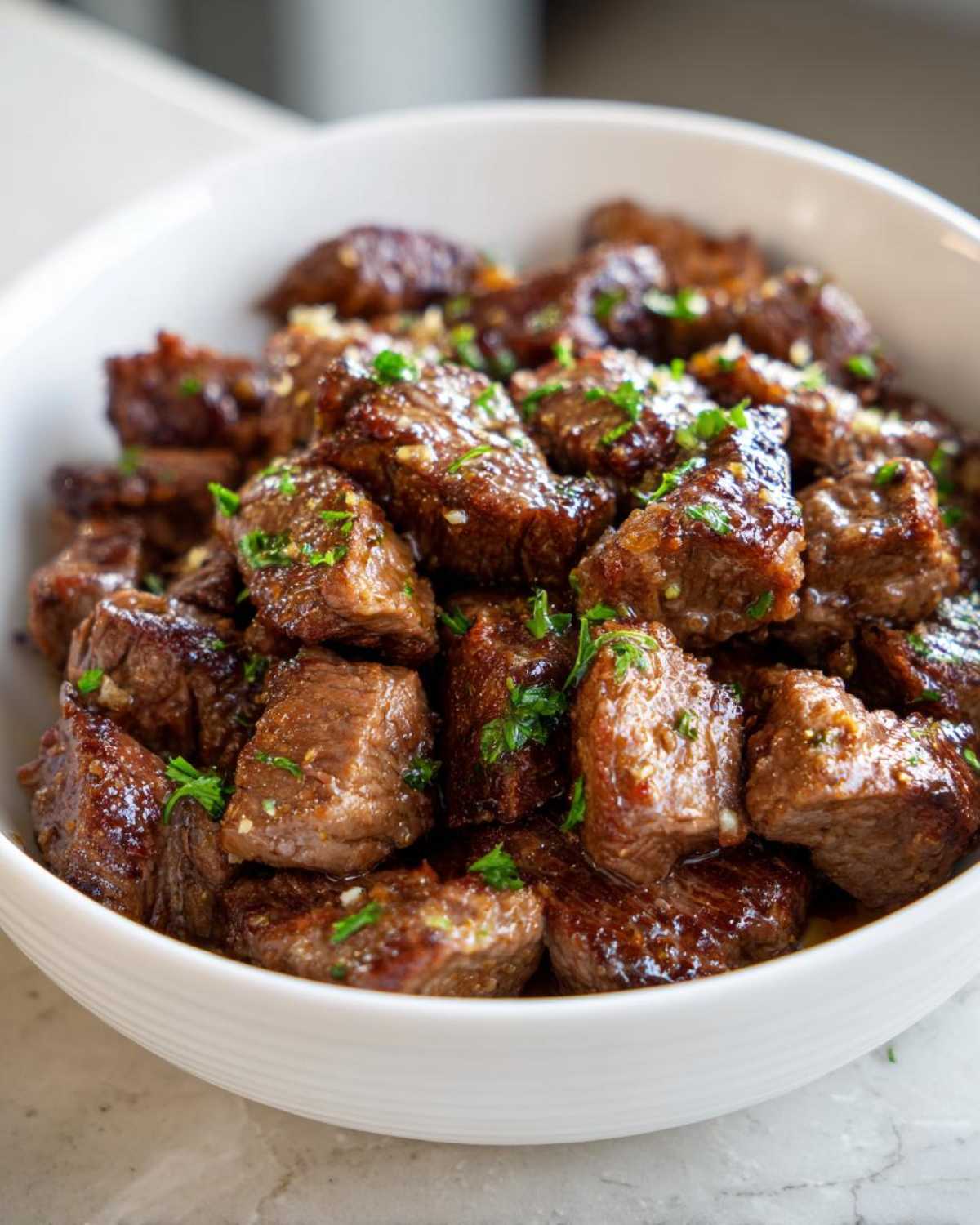 Close-up of tender, glazed crockpot steak bites served in a white bowl and sprinkled with fresh parsley.