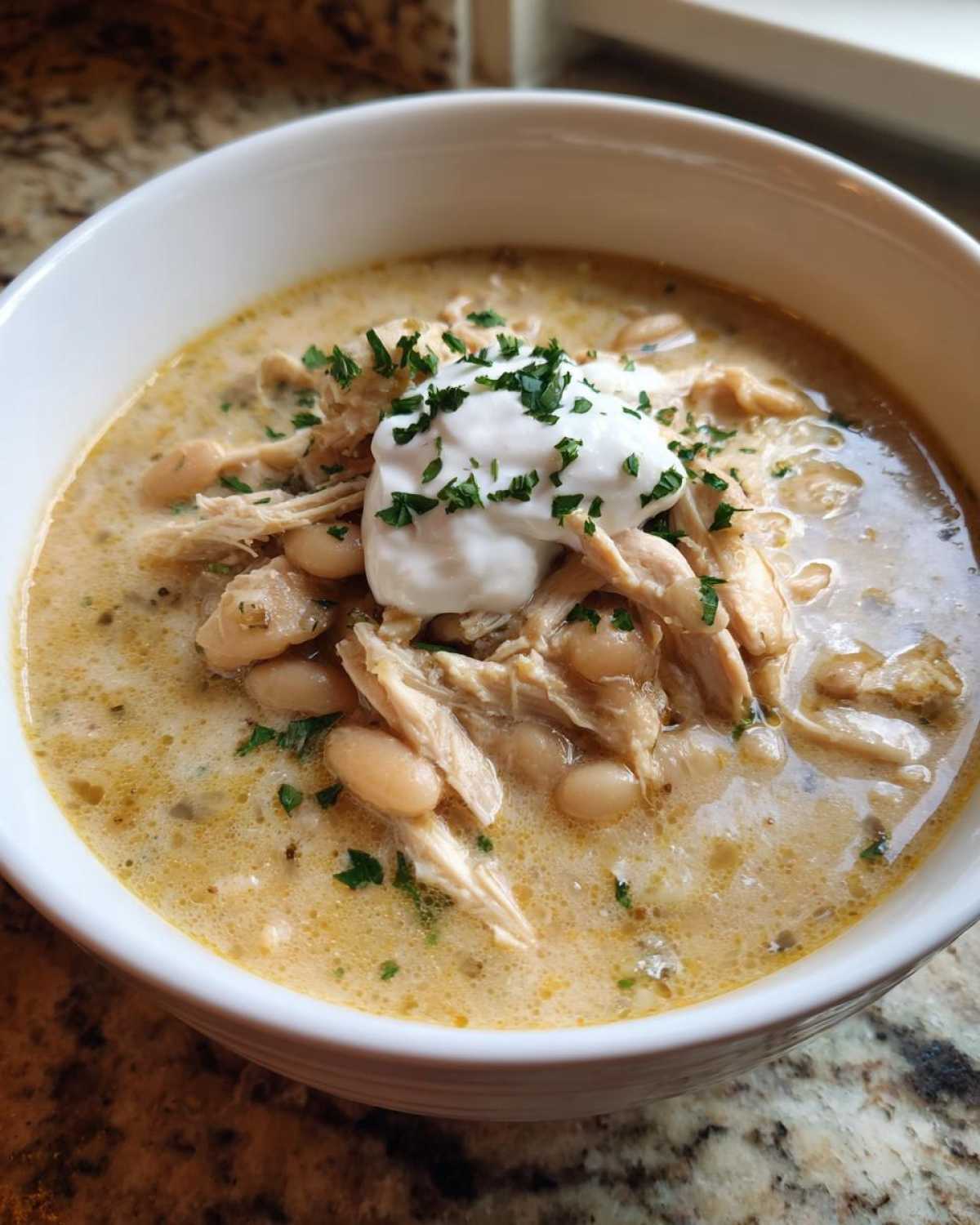 A close-up of a bowl of creamy crockpot white chicken chili topped with sour cream and fresh parsley.
