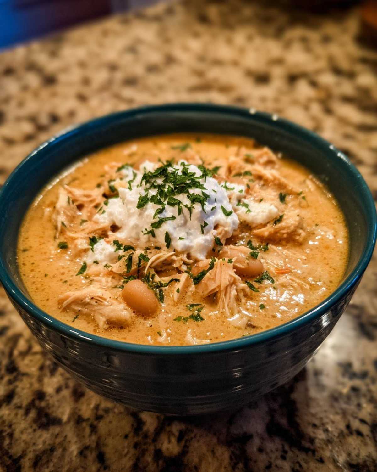 A close-up of a bowl of creamy crockpot white chicken chili topped with sour cream and parsley.