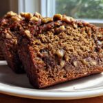 Three thick slices of moist date nut bread, topped with walnuts, displayed on a white plate.
