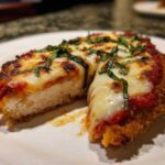 A close-up view of a slice of Easy Chicken Parmesan showing the breaded exterior, thick white chicken breast, melted mozzarella, tomato sauce, and fresh basil.