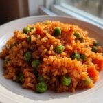 A close-up of a serving of bright orange Spanish rice mixed with green peas and diced carrots on a white plate.