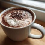 Close-up of a steaming white mug filled with rich hot cocoa mix, topped with foam and chocolate shavings.