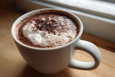 Close-up of a steaming white mug filled with rich hot cocoa mix, topped with foam and chocolate shavings.