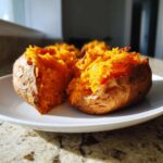 A fluffy baked sweet potato, split open to show the bright orange interior, served on a white plate.