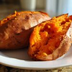 Close-up of a fluffy baked sweet potato split open, revealing bright orange flesh on a white plate.