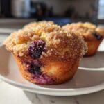Close-up of a fluffy blueberry muffin topped with sparkling coarse sugar, sitting on a white plate.