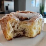 A close-up shot of a fluffy, freshly baked cinnamon roll with a bite taken out, covered in white icing.