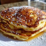 A close-up of a tall stack of fluffy sourdough pancakes drizzled with syrup and dusted with powdered sugar.