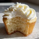 Close-up of a vanilla cupcakes with a bite taken out, showing the fluffy crumb and swirl of white frosting.