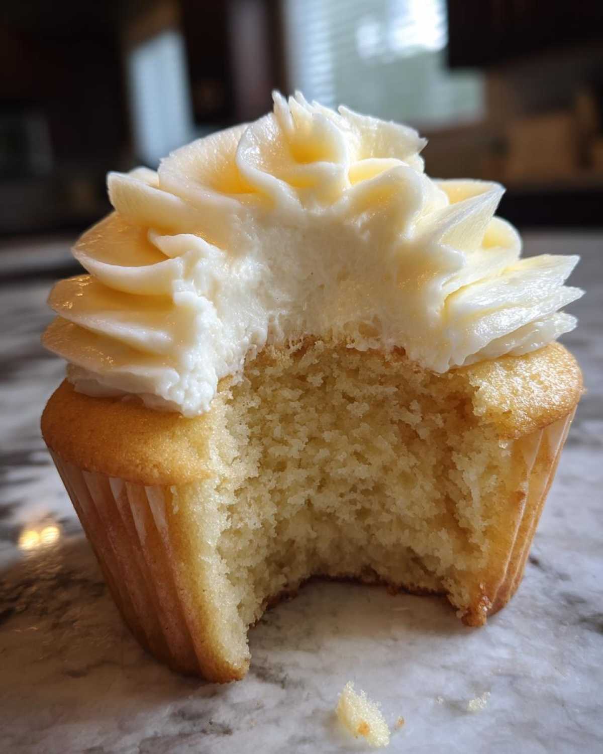 A close-up of a vanilla cupcake with a bite taken out, revealing the fluffy interior and thick swirl of white vanilla frosting.