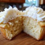 Cross-section of a fluffy vanilla cupcake showing the moist crumb and thick swirl of white frosting.
