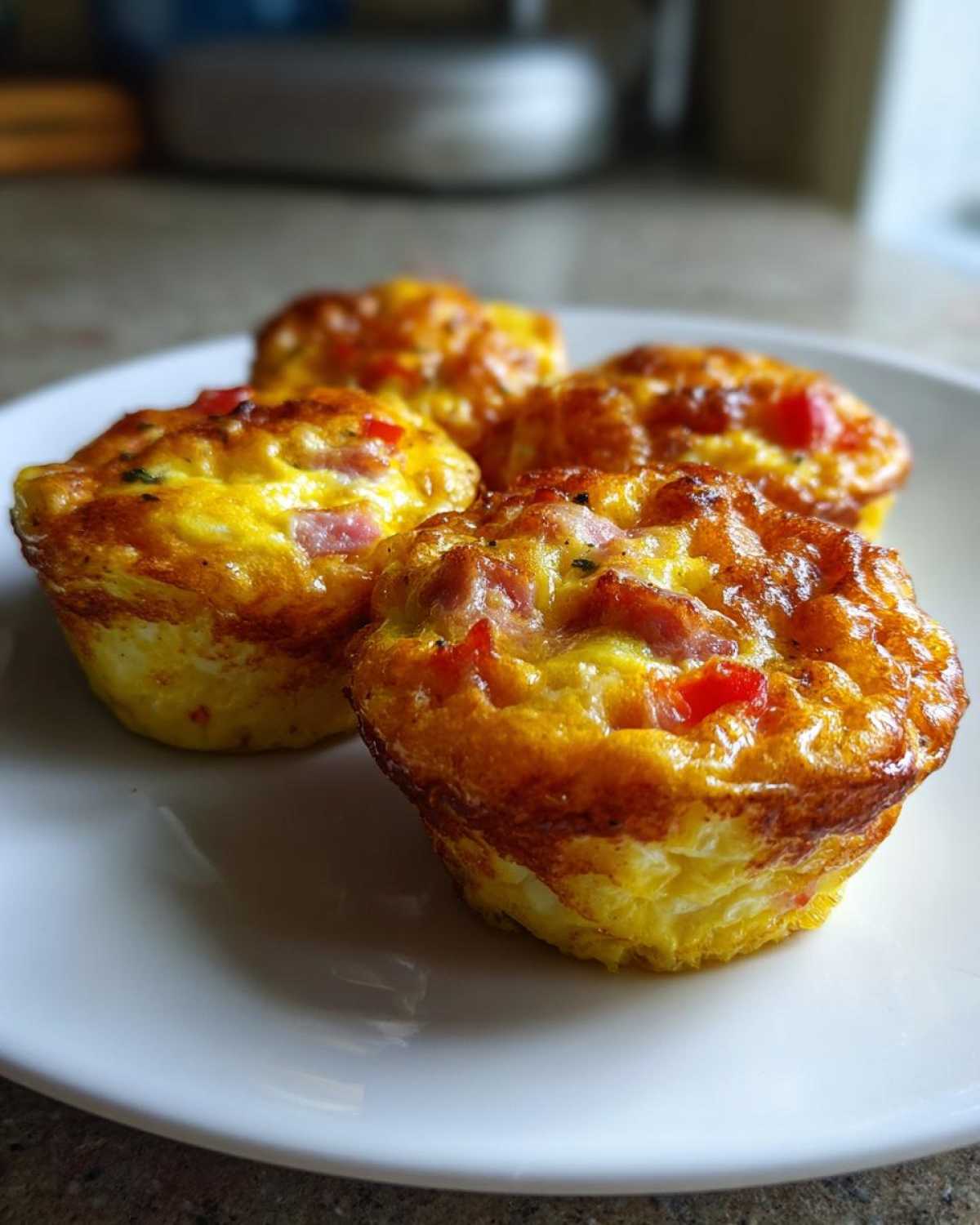 Four freshly baked Egg Muffin Cups with visible ham and red pepper pieces, sitting on a white plate.
