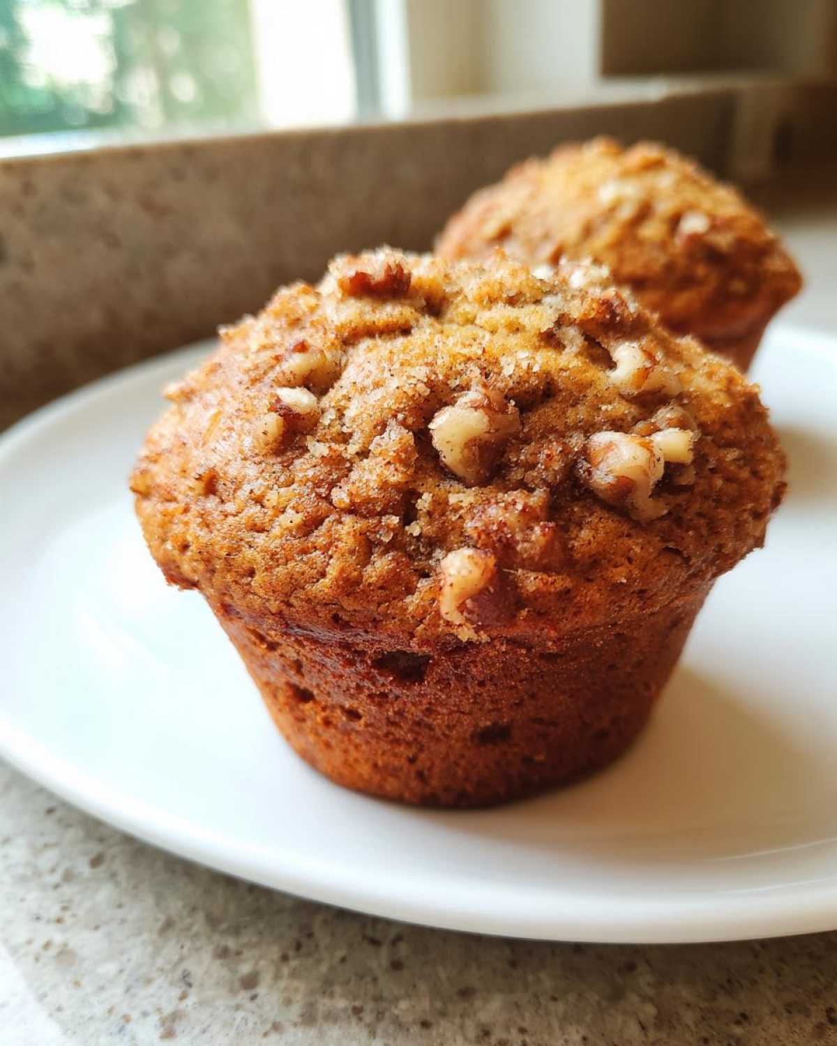 A close-up of a freshly baked banana nut muffin topped with walnuts and cinnamon sugar, resting on a white plate.