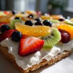 Close-up of a rectangular fruit pizza sugar cookie topped with white frosting, strawberries, kiwi, oranges, and blueberries.