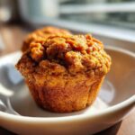 A close-up of a freshly baked, golden brown honey bran muffin with a crumbly top, resting on a white dish.