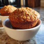 A close-up, sunlit photo of a golden brown honey bran muffin sitting in a small white bowl.