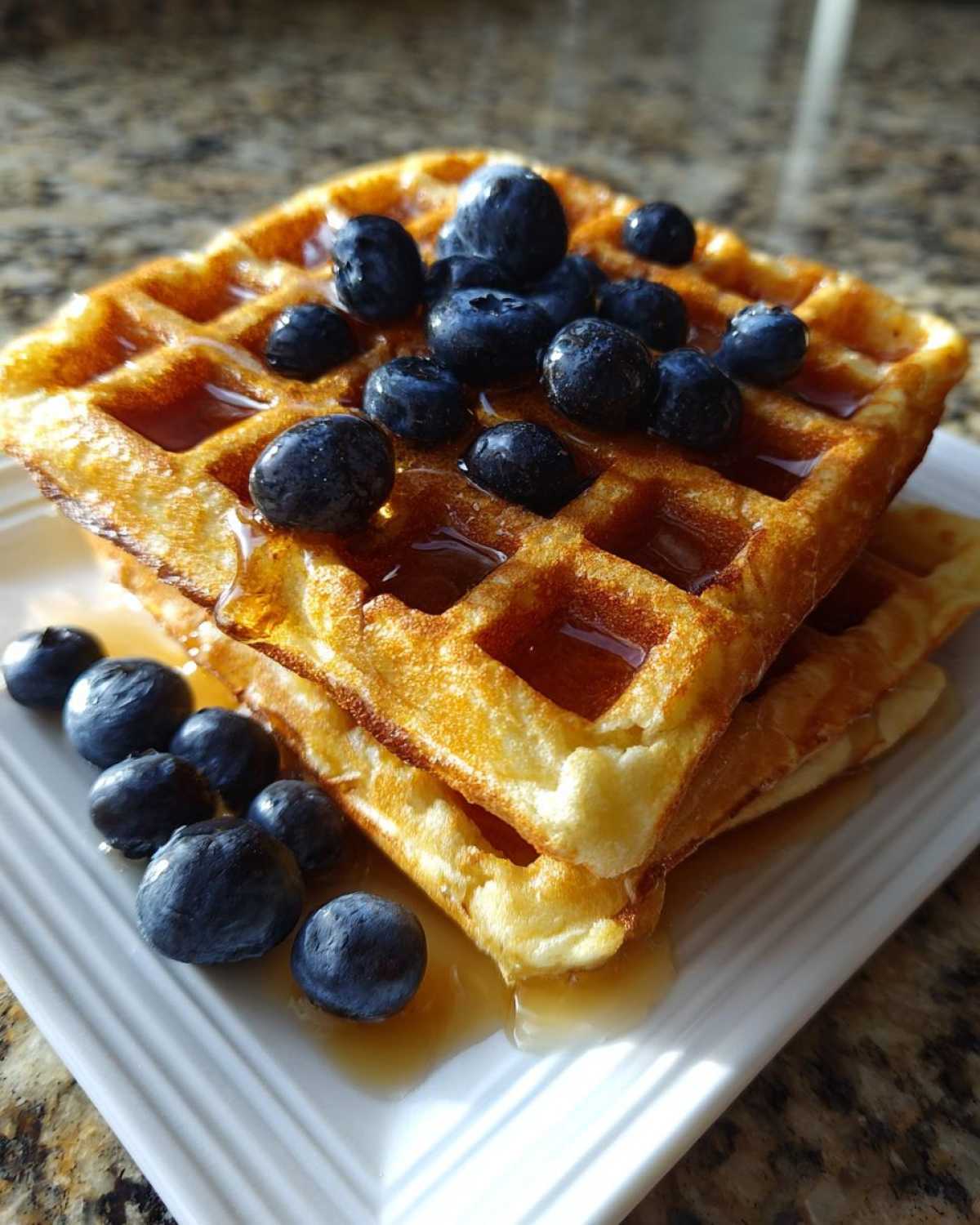 Stack of golden brown high protein waffles topped with fresh blueberries and drizzled with syrup.