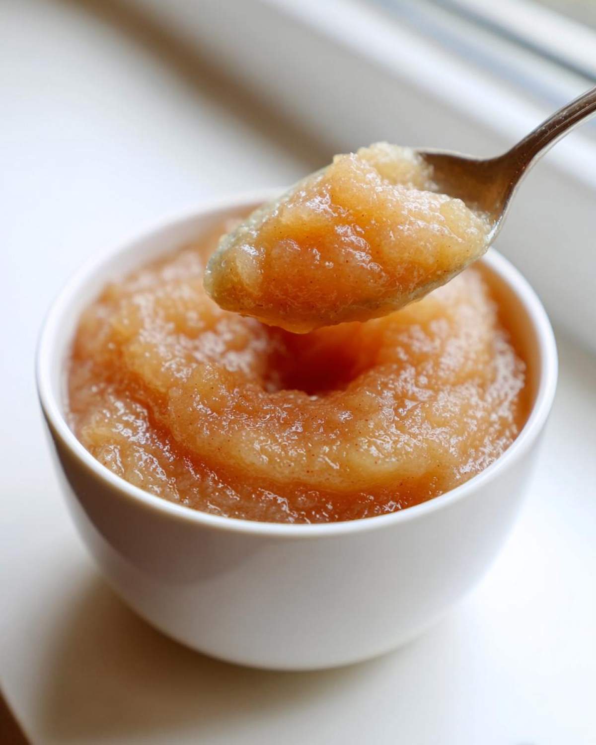 A spoonful of chunky, homemade applesauce being lifted from a small white bowl.