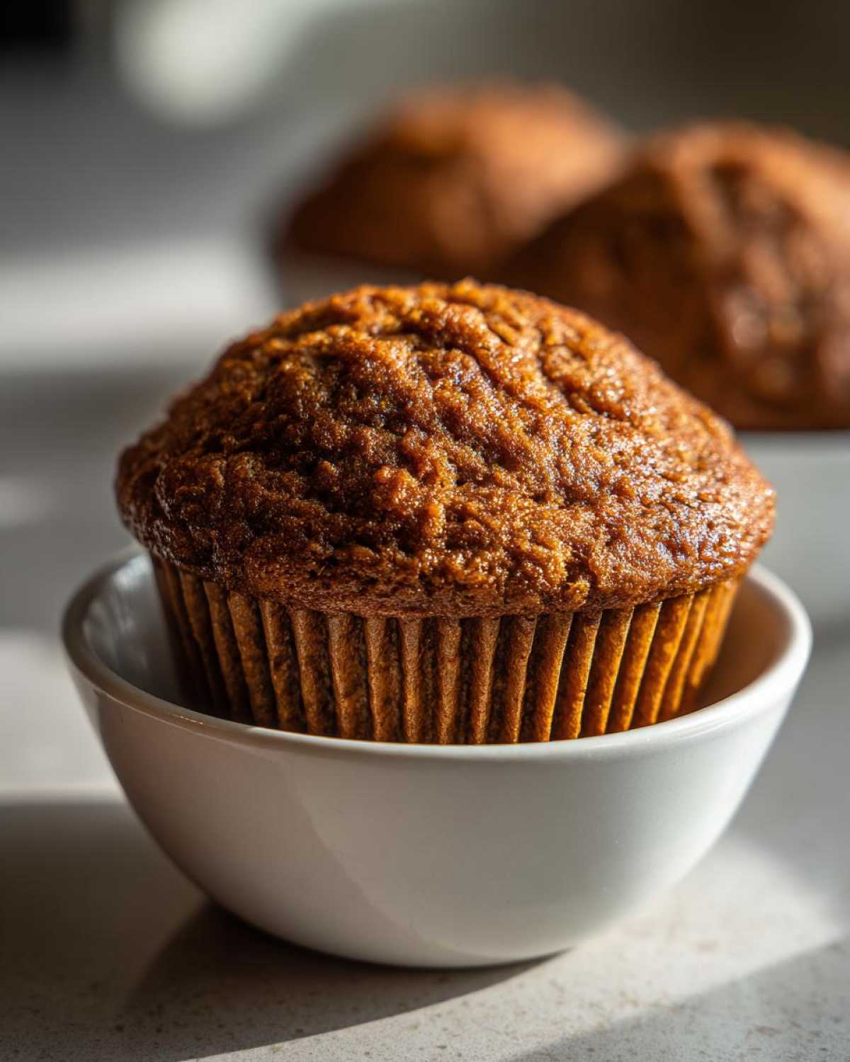 Close-up of a richly browned honey bran muffin sitting in a small white bowl with more muffins blurred in the background.