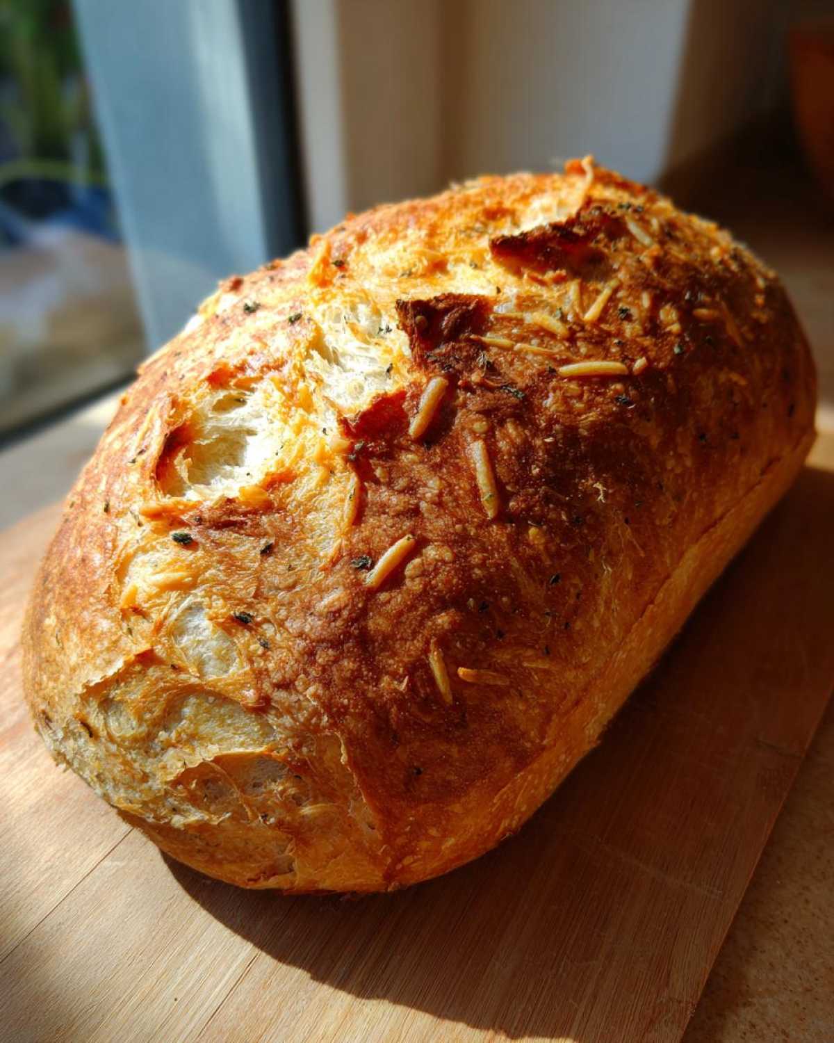 A freshly baked, golden-brown loaf of Italian herb and cheese bread resting on a wooden cutting board.