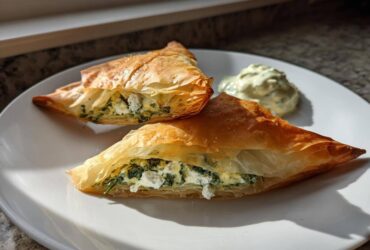 Two golden, flaky Mediterranean Spinach and Feta Cheese Crisps triangles served with a dollop of creamy dip.