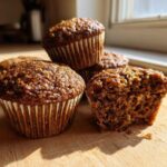 A stack of moist bran muffins on a wooden board, with one muffin broken open showing the texture.