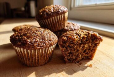 A stack of moist bran muffins on a wooden board, with one muffin broken open showing the texture.