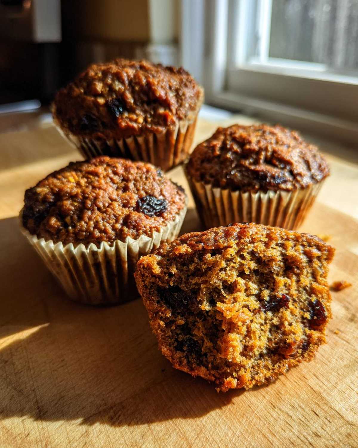 Four moist bran muffins, one broken open to show the texture and raisins inside, sitting on a wooden board.
