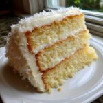A thick, moist slice of three-layer coconut cake with white frosting and shredded coconut topping.