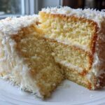 Cross-section of a moist coconut cake slice showing two layers, white frosting, and covered in shredded coconut.