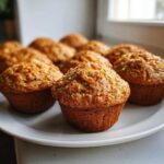 A plate holding several freshly baked morning glory muffins, showing visible shreds of carrot and nuts on top.