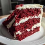 Close-up of a tall, moist slice of red velvet cake showing three layers of deep red cake and thick white cream cheese frosting.