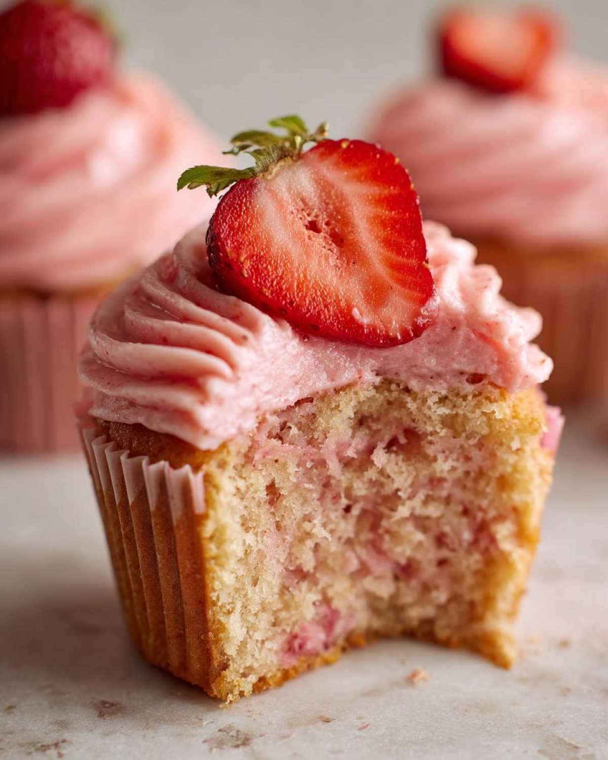 Close-up of a strawberry cupcake with a bite taken out, showing the moist crumb and pink frosting topped with a strawberry slice.