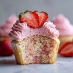 Close-up of a strawberry cupcake cut in half showing moist cake and pink frosting, topped with fresh strawberry slices.