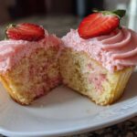 Cross-section view of moist strawberry cupcakes showing pink frosting and fresh strawberry slices on top.