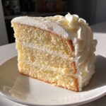 Close-up of a moist slice of layered vanilla cake recipe with thick white buttercream frosting.