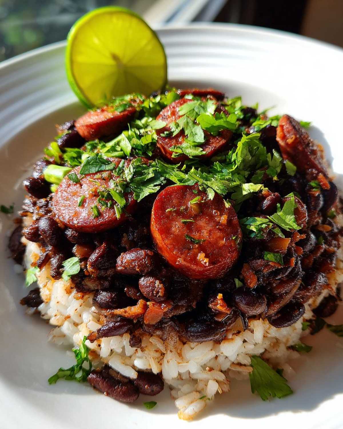 A close-up of flavorful black beans and rice topped with sliced sausage and fresh cilantro, garnished with a lime wedge.