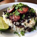 A close-up of flavorful black beans and rice topped with seared sausage slices and fresh cilantro, served with a lime wedge.