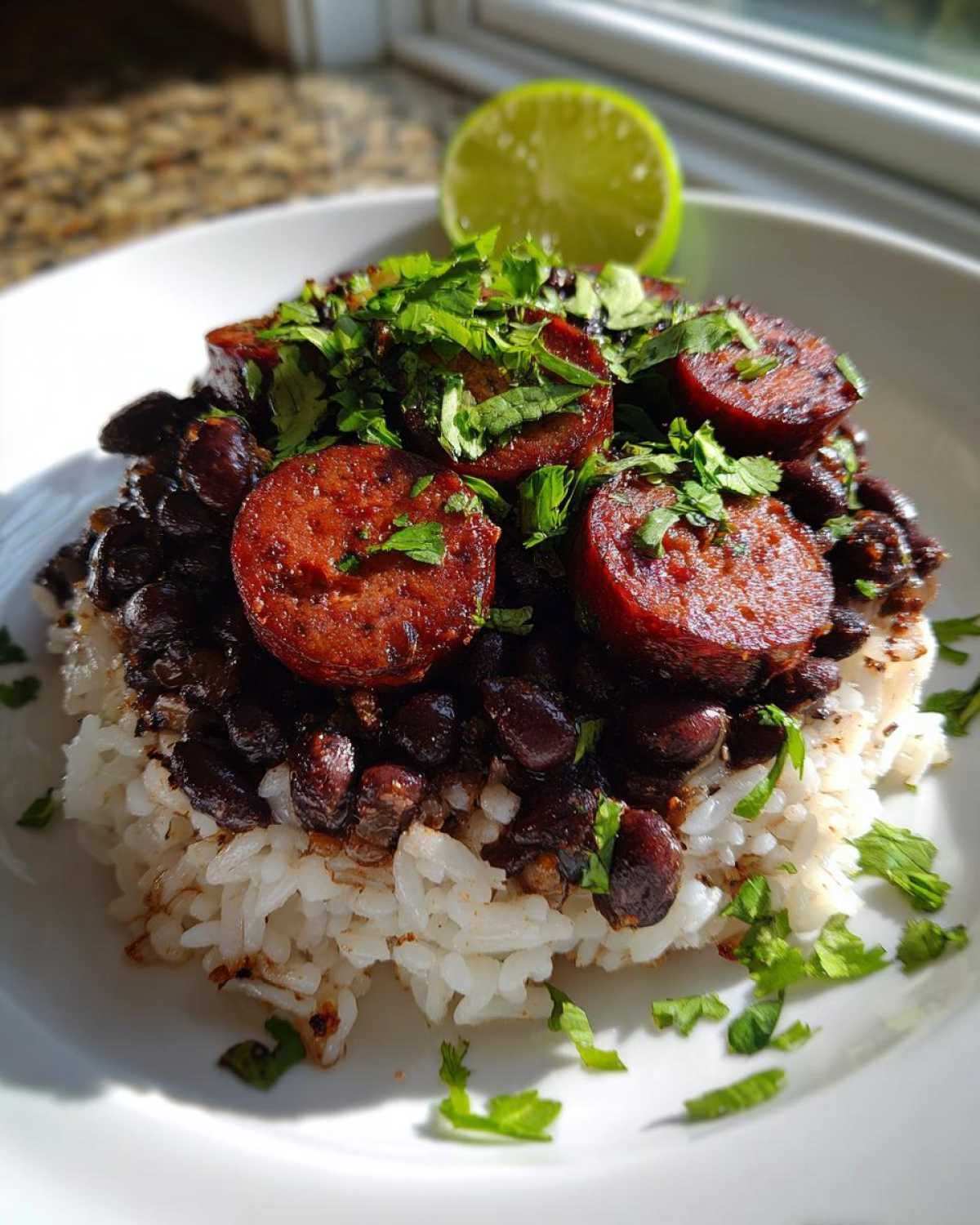 A close-up of a serving of black beans and rice topped with sliced sausage, cilantro, and a lime wedge.