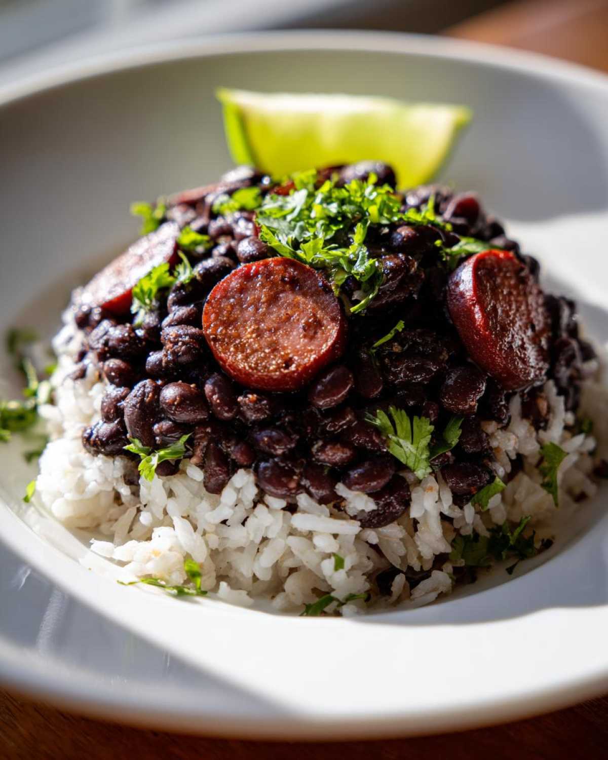A serving of flavorful black beans and rice topped with sliced sausage and fresh cilantro, served with a lime wedge.