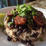 A close-up serving of flavorful black beans and rice topped with sliced sausage and fresh cilantro, with a lime wedge on the side.