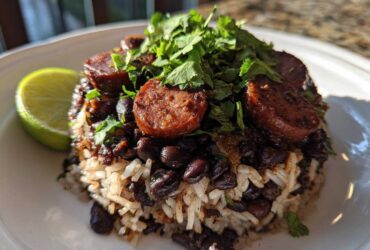 A close-up serving of flavorful black beans and rice topped with sliced sausage and fresh cilantro, with a lime wedge on the side.