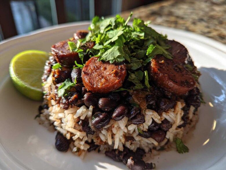 A close-up serving of flavorful black beans and rice topped with sliced sausage and fresh cilantro, with a lime wedge on the side.