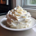 A generous swirl of fluffy, white cream cheese frosting piped high on a small white plate.