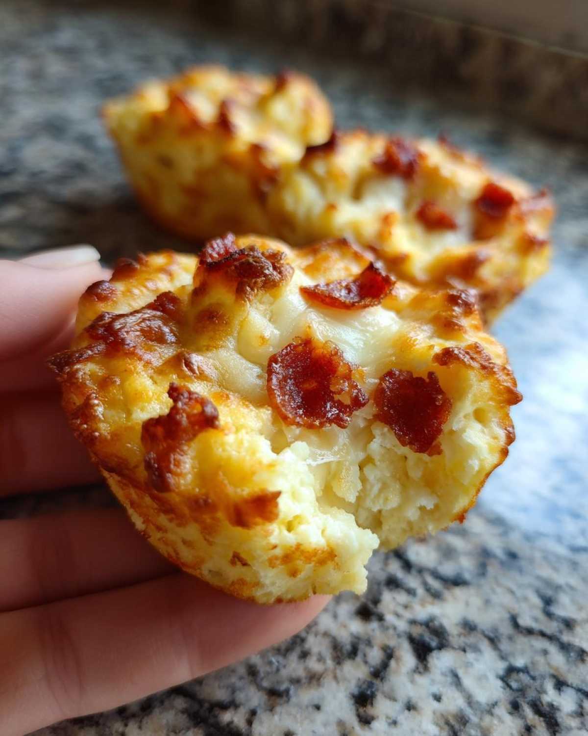 A hand holding a freshly baked pizza muffin with melted cheese and pepperoni, showing a bite taken out.