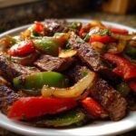 Close-up of sizzling steak fajitas featuring sliced beef, red and green bell peppers, and onions on a white plate.