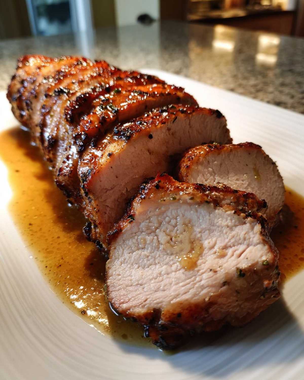 Close-up of a juicy, sliced pork loin roast resting in its pan juices on a white plate.